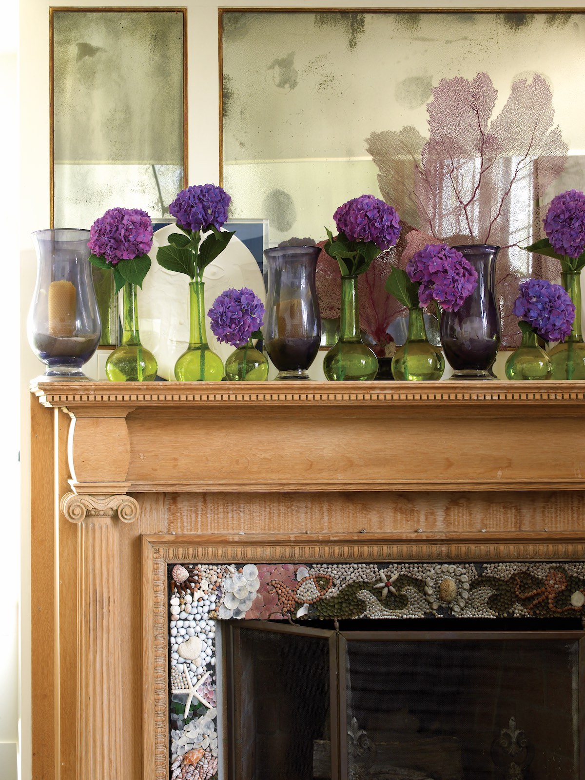 Carved wooden mantle topped with stems of purple hydrangeas in green glass bud vases.