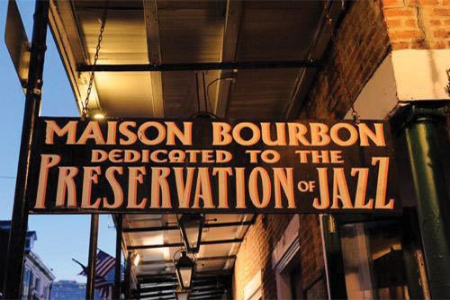 A sign hanging from a traditional New Orleans balcony reads "Maison Bourbon: Dedicated to the Preservation of Jazz"