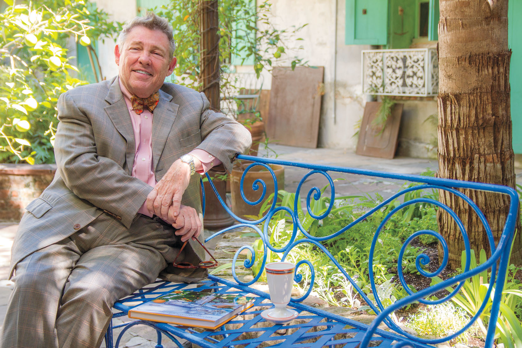 Portrait of New Orleans interior decorator and antiques shop owner Patrick Dunne, wearing a gray suit with pink shirt, sitting outside a wrought-iron bench painted bright blue