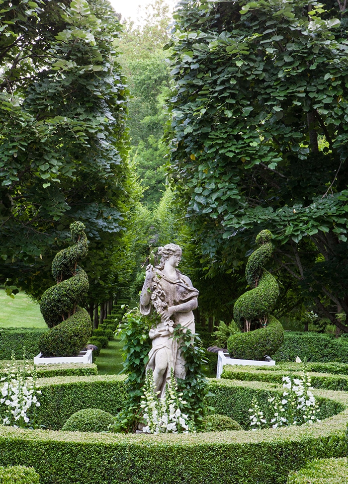 Carolyne Roehm garden parterre: In a precisely pruned shrubbery circle, lush vines climb a Classical-style statue of a woman, which stands in a bed of white foxglove flowers. In the background, two swirling topiaries mark the entrance to tree-lined path.