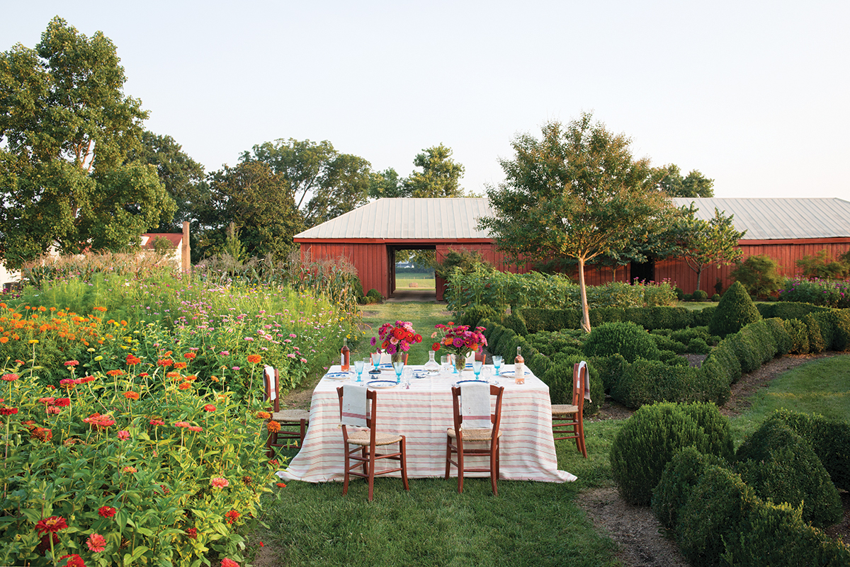 Table for six, set in a field of zinnias with a red barn in the background. The table is set with red and white linens, blue and white china, and two vases of pink and red zinnias.