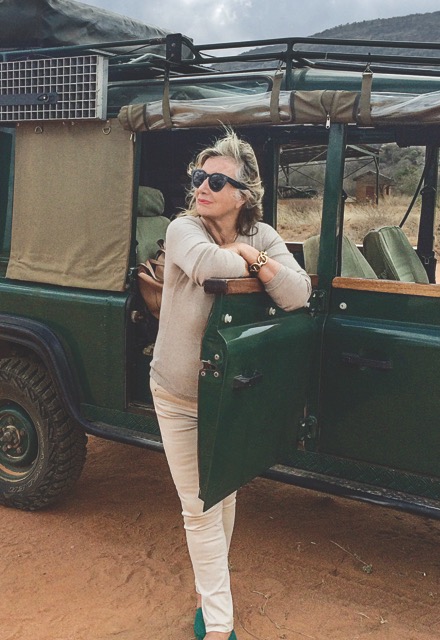 Sylvie Chantecaille stands by a green truck in the Africian countryside, wearing sunglasses, and a beige sweater, light pants, and deep turquoise flats