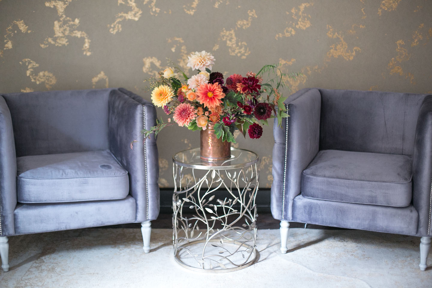 A floral arrangement in a copper vessel stands on a small metal drinks table between two blue-gray velvet, square-shaped wing chairs. The wallpaper is gray with a metallic gold marbling.