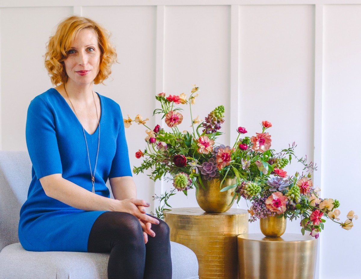 portrait of interior designer Shay Geyer wearing a royal blue blouse, posing with her arms crossed. A vase of blue and white hydrangeas and a wallpaper featuring a pattern of undulating blue lines are in the background