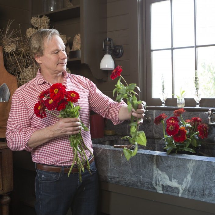 P. Allen Smith, wearing a red gingham shirt and jeans, stands at a farmhouse-style sink with bunches of red zinnias