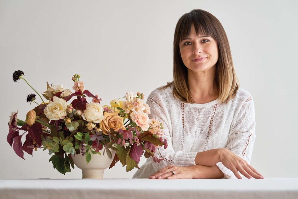 portrait of flora designer Adri Smith of Moss Floral, wearing a white top and shoulder length hair with bangs. She leans on a white counter next to her finished arrangement, with her arms resting crossed on the counter surface