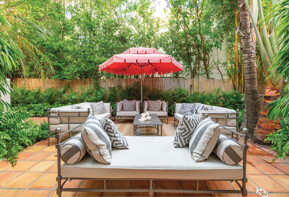 Photo of a terra cotta tile patio with outdoor seating surrounding a coffee table shaded by a red umbrella. Gray-and-white throw pillows feature stripes and a Greek key pattern.