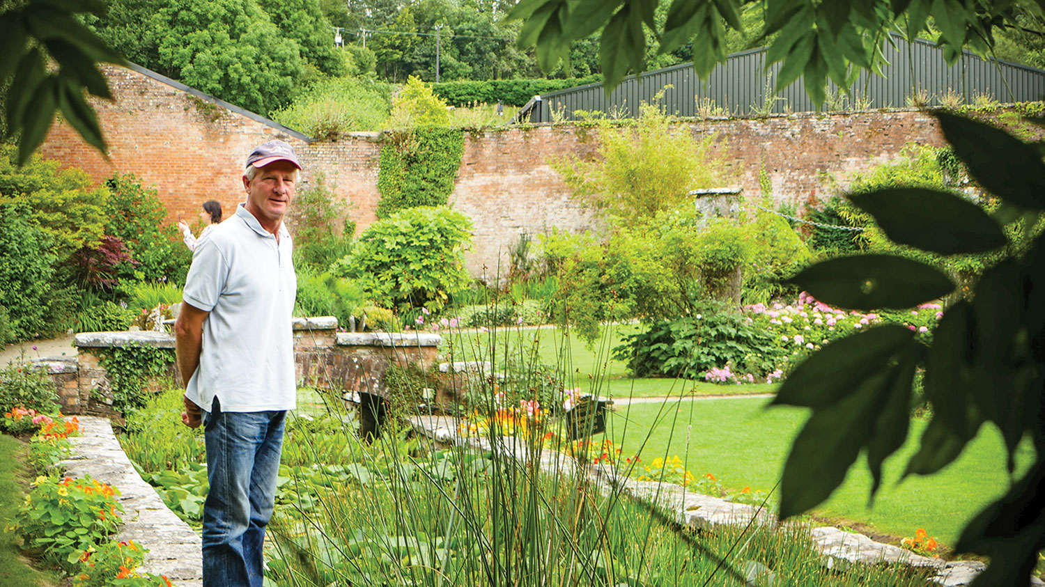 Wearing a baseball cap, polo shirt and jeans, Arthur Shackleton stands on the stone edge of a plant filled garden pond, holding his hands behind his back.