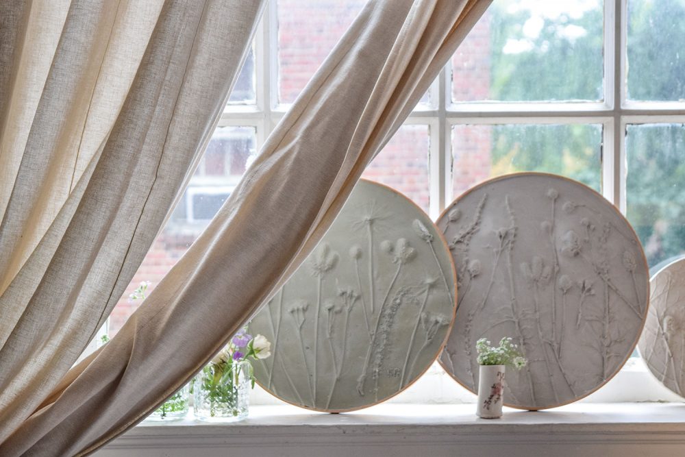 Three circular Ron Nicole pieces stand on a window sill with a college of bouquets in small porcelain and cut-glass vases. There's a muslin curtain tied back, and the window is an older style with grilles.