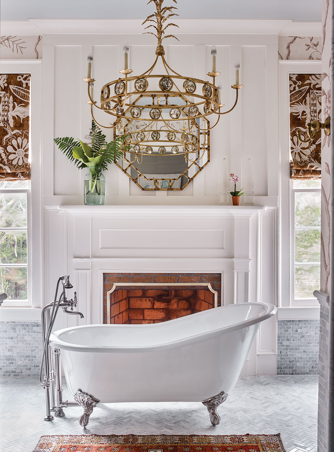 A photo of a master bath designed by James Farmer featuring a clawfoot tub under a gilded chandelier, in front of a fireplace, and light gray herringbone-pattern tile floor.