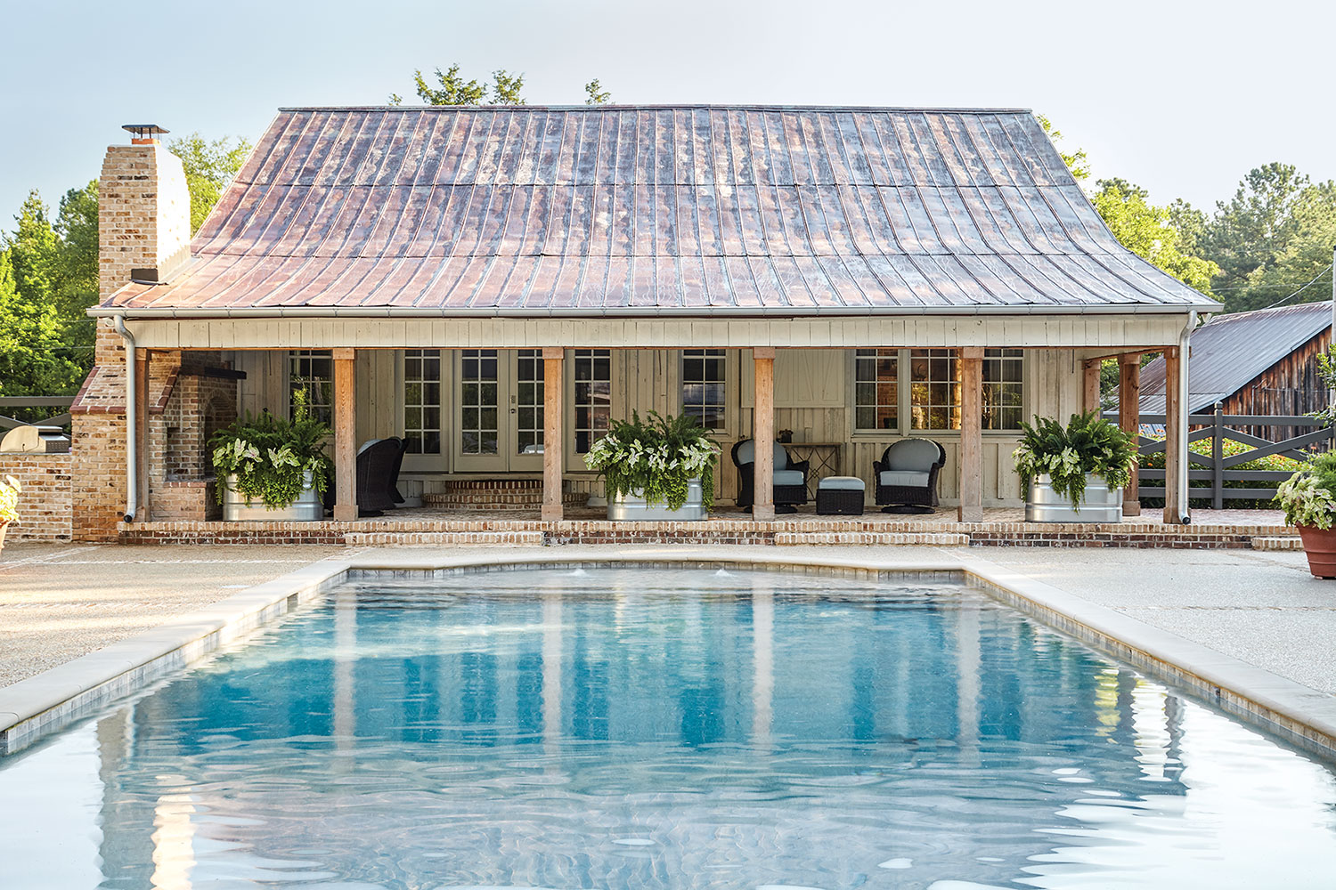 A blue pool leads to a covered brick patio of the pool house, which features wood columns across the front and an aged metal roof and brick chimney