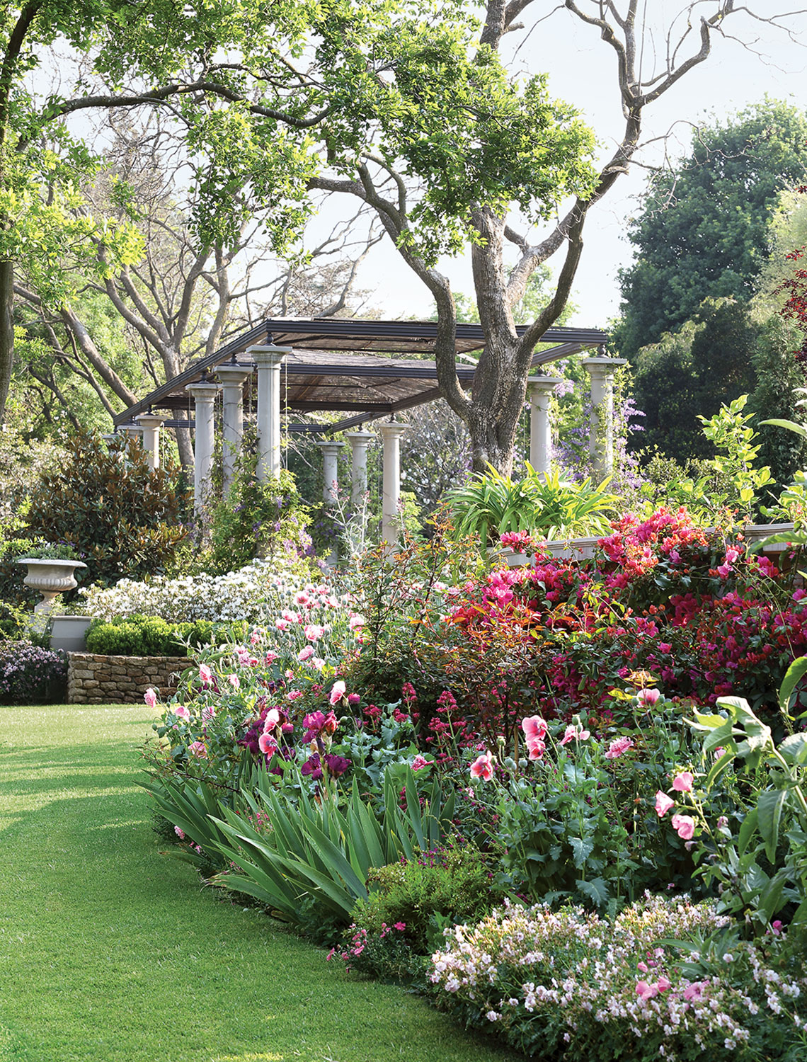 Various pink flowers in foreground and trees and a pergola in the background