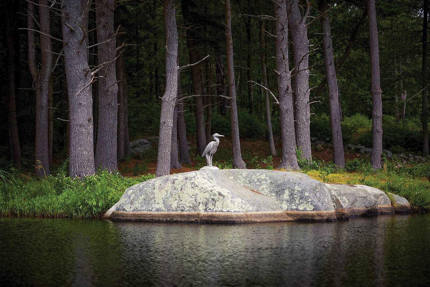 The statue stands on a large rock at the edge of a body of water. In the background is a dense forest.