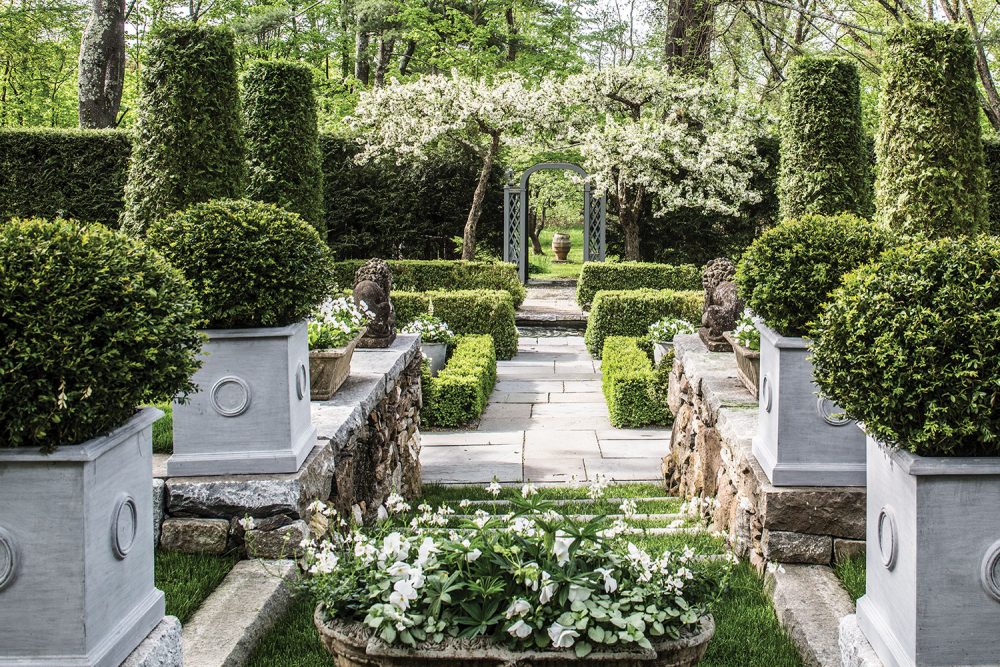 View through the center path of a formal garden filled with shrubs trimmed into geometric shapes, such as spheres, columns and square boxwood borders. The Pennoyer Newman Circle Icon Planters are square and several feet high and wide. In the distance, you can see an arbor leading to the exit, which is framed by a small flowering tree or shrub.