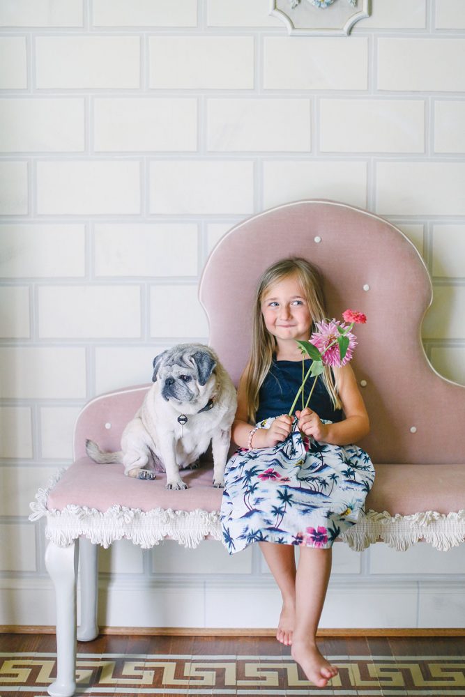 A little girl, who holds fresh flowers in each hand, sits on a pink upholstered bench beside a pug in Jason Oliver Nixon and John Loecke's home in High Point, North Carolina