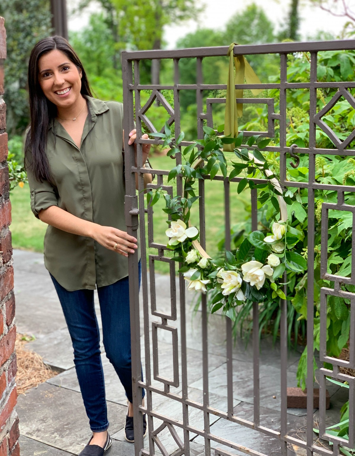 Jessica Cohen stands at a wrought iron gate decorated with a floral hoop of gardenias, spray roses, and Italian ruscus