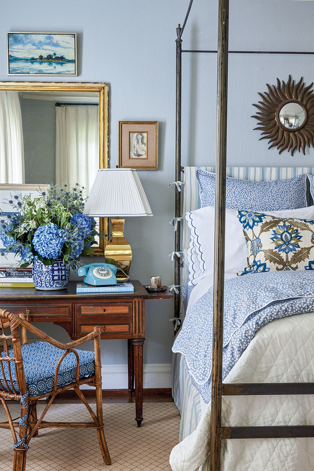 A desk and chair beside a four-poster bed is decorated with a large arrangement of hydrangeas, a vintage-inspired blue phone with rotary dial, a brass lamp, and large rectangular mirror with a brass frame. The walls are painted a pale blue