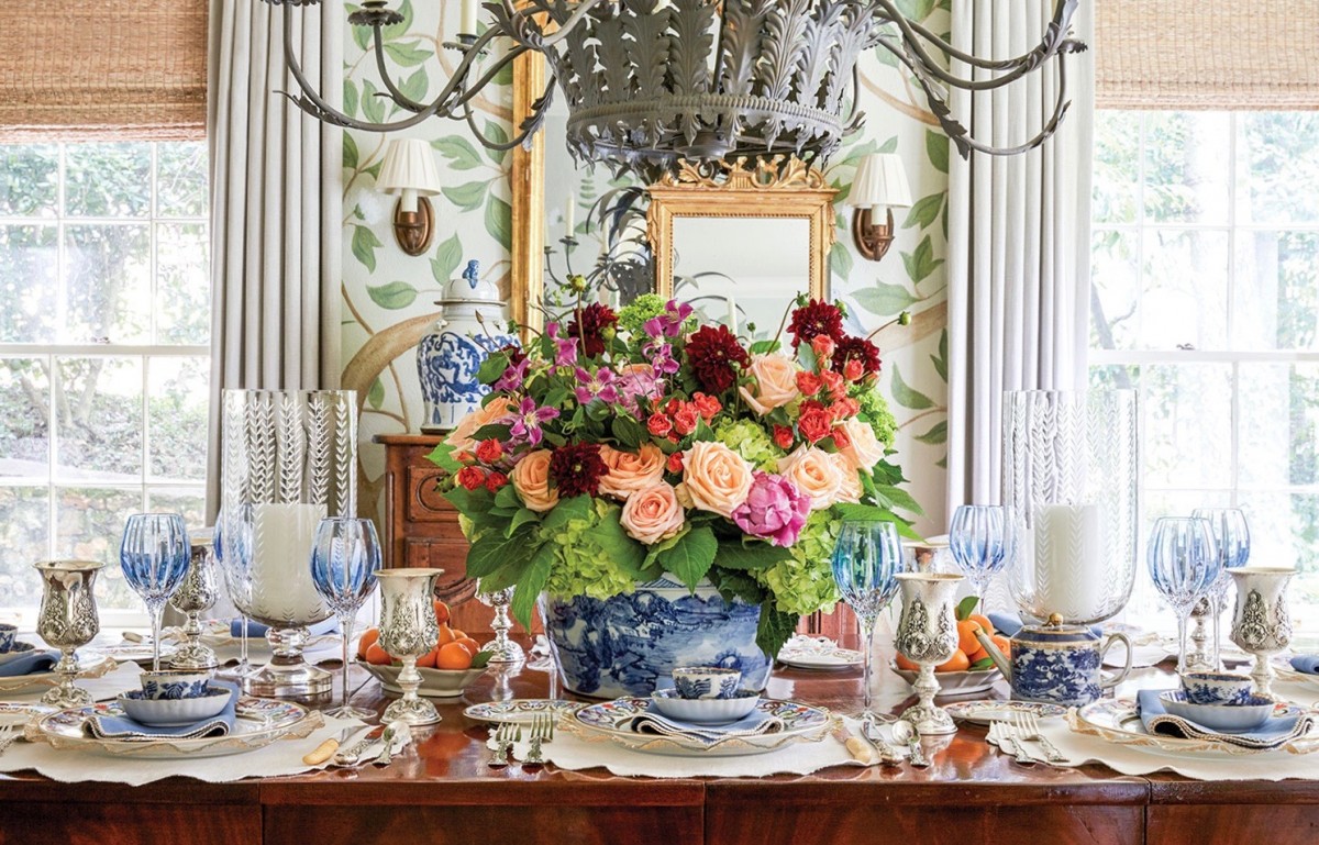 A large wooden table graciously set with blue-and-white china, blue-tinted crystal goblets, silver goblets, two large glass hurricanes holding a large candle each, and—at the center—a large blue-and-white china tureen filled with a flower arrangement of peach, pink, green, coral, and burgundy blooms.