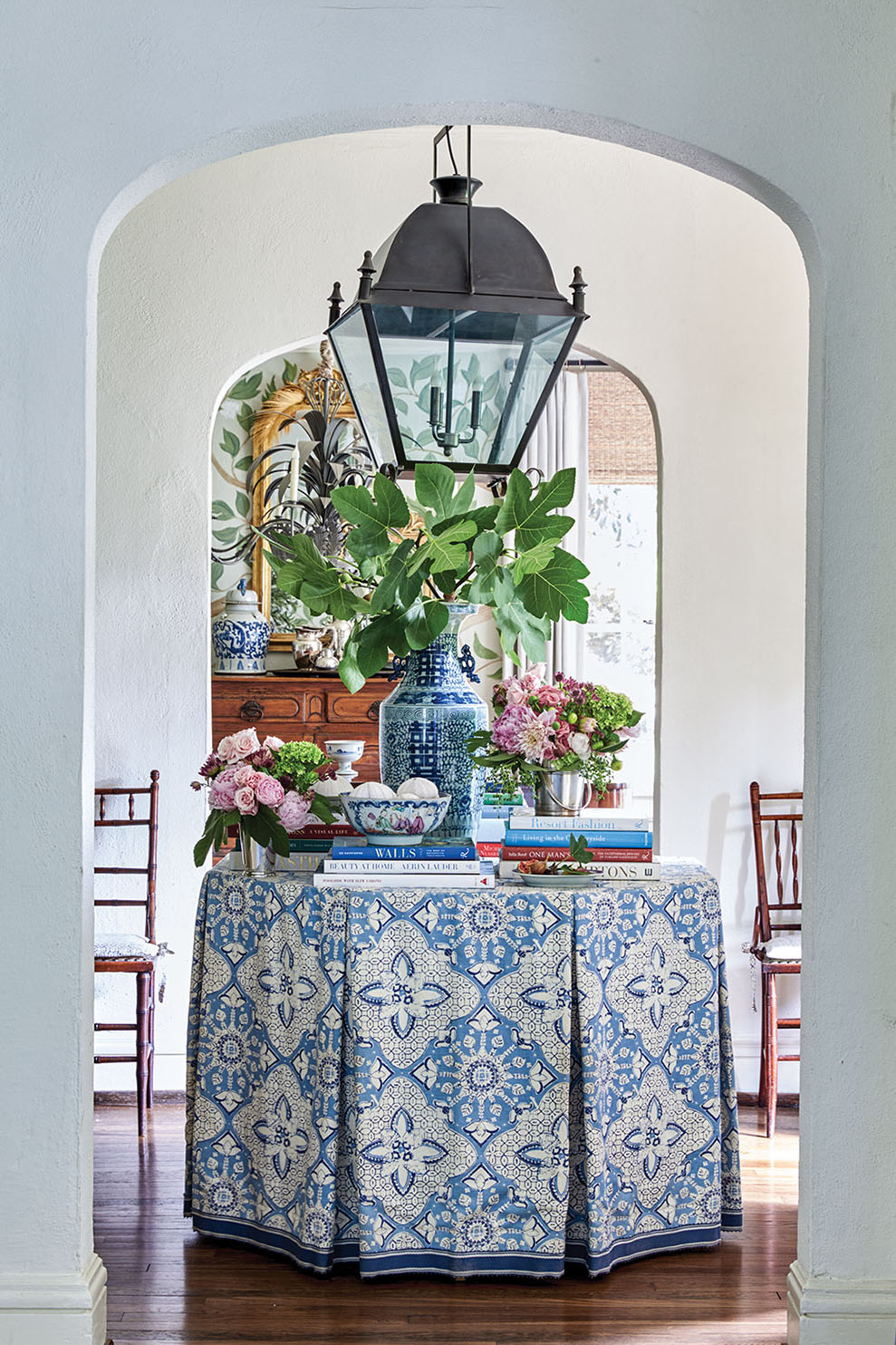 An octagonal table in an entry is topped with a tailored blue-and-white table skirt, blue-and-white pottery, books, and floral arrangements.
