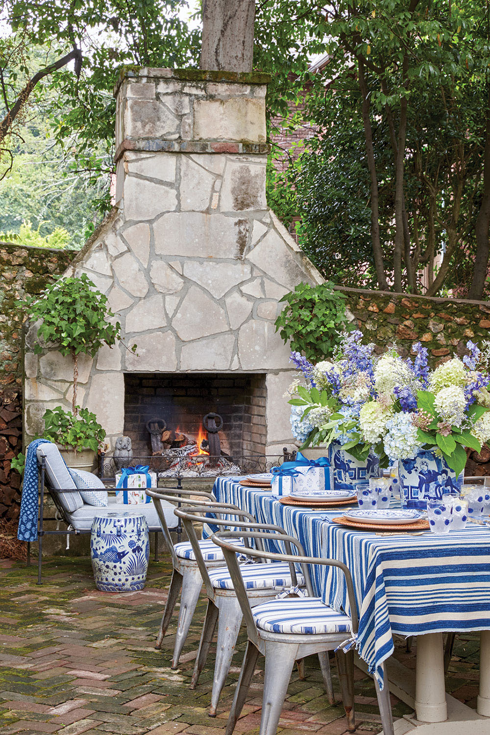 Another view of the outdoor table shows metal dining chairs with blue-and-white striped seats and an outdoor stone fireplace with a roaring fire.