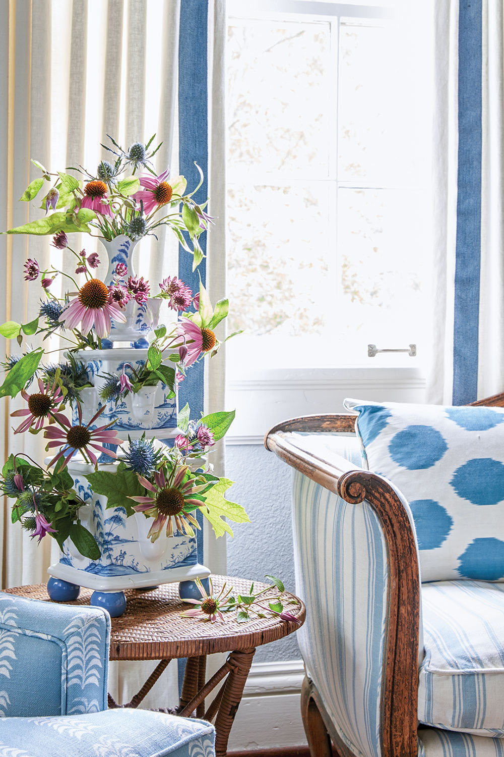 The flower arrangement sits on a side table in the bedrooms sitting area between a settee and a chair, both upholstered in various patterns of blue-and-white, including stripes, dots, and a botanical leaf print