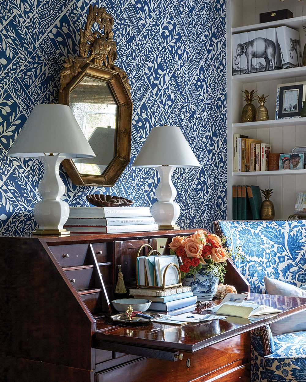 A blue-and-white wallpapered room with a matching chair, a wooden writing desk with drop-down top, two white lamps, a gilded mirror, and white built-in bookshelves.