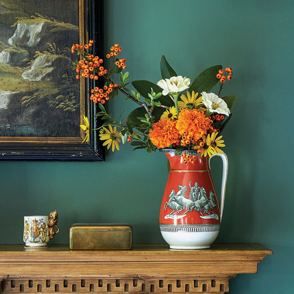 Red and white pitcher with arrangement of marigolds, zinnias, black-eyed Susans, and pyracantha berries on wooden mantel.