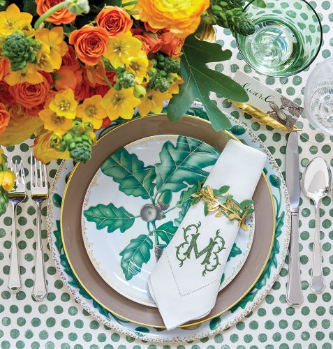 Fall table setting featuring green accents, including a leaf motif on the plates, the monogram on the napkin, and polka dots on the tablecloth. A flower arrangement of yellow and orange contrasts with the green, allowing all the colors to pop.