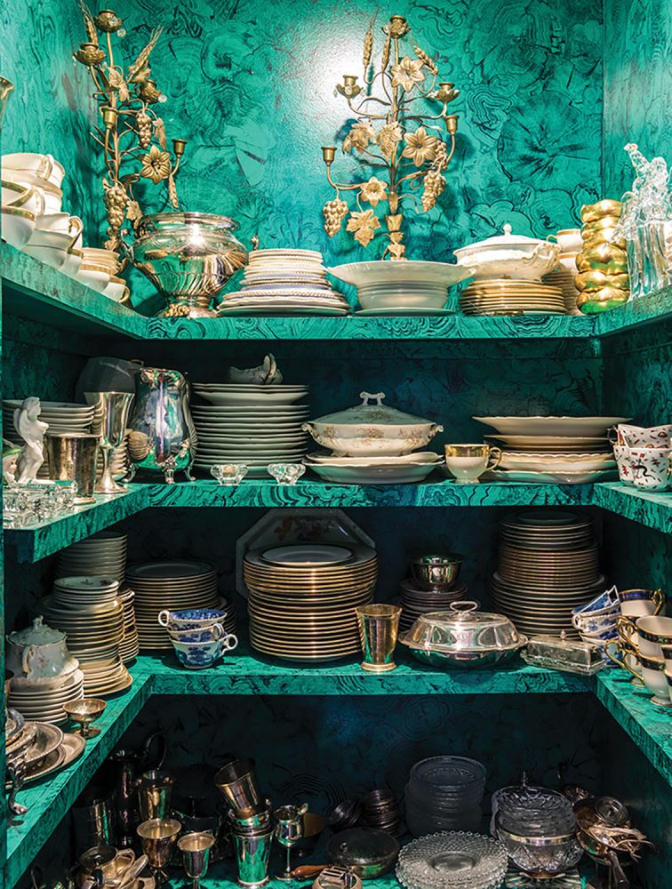 A china closet with four levels of shelves of three sides holds stacks of china, elegant gilt candelabras, and silver serving pieces. Bright teal walls and matching shelves feature a pattern that resembles marbling.