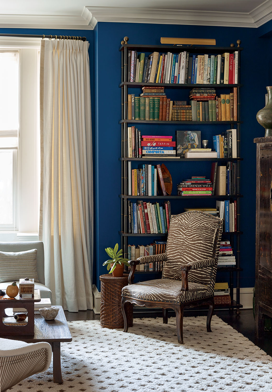 A zebra-print upholstered armchair punctuates the sitting area of a Tom Scheerer-designed room. Natural white, floor-to-ceiling curtain panels and a plush natural white rug, which features a raised pattern outlining diamond shapes, balances the deep blue paint on the walls. A tall, full set of bookshelves gives the room a lived-in look.