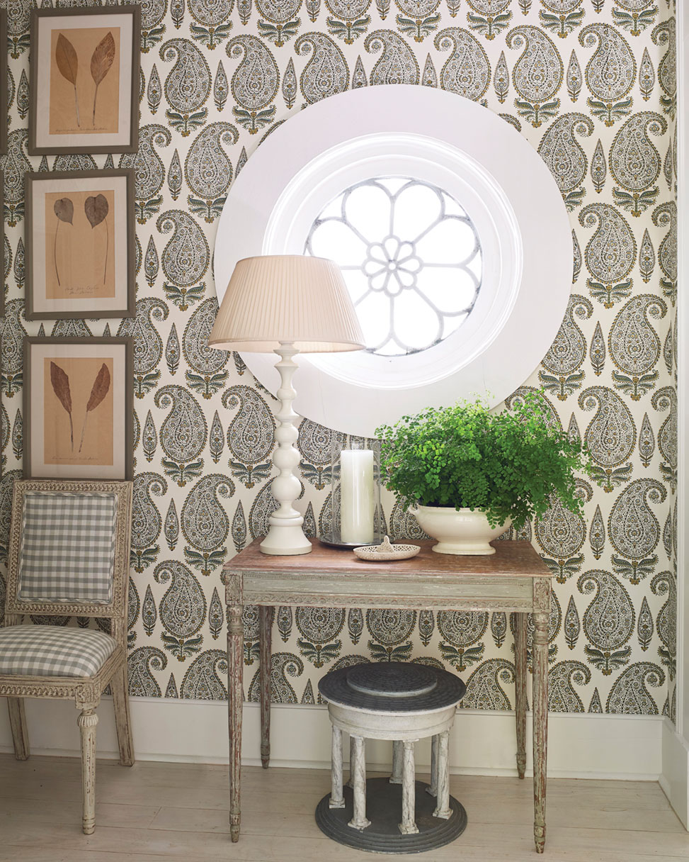 The Cathy Kincaid-designed room features an all-neutral palette, with a green fern adding a pop of color. A small table and lamp sit beneath a circular leaded window with a floral motif. A small chair is upholstered in a khaki and white check, contrasting with the paisley wallpaper. Above the chair, three leaf prints hang in a vertical column configuration.