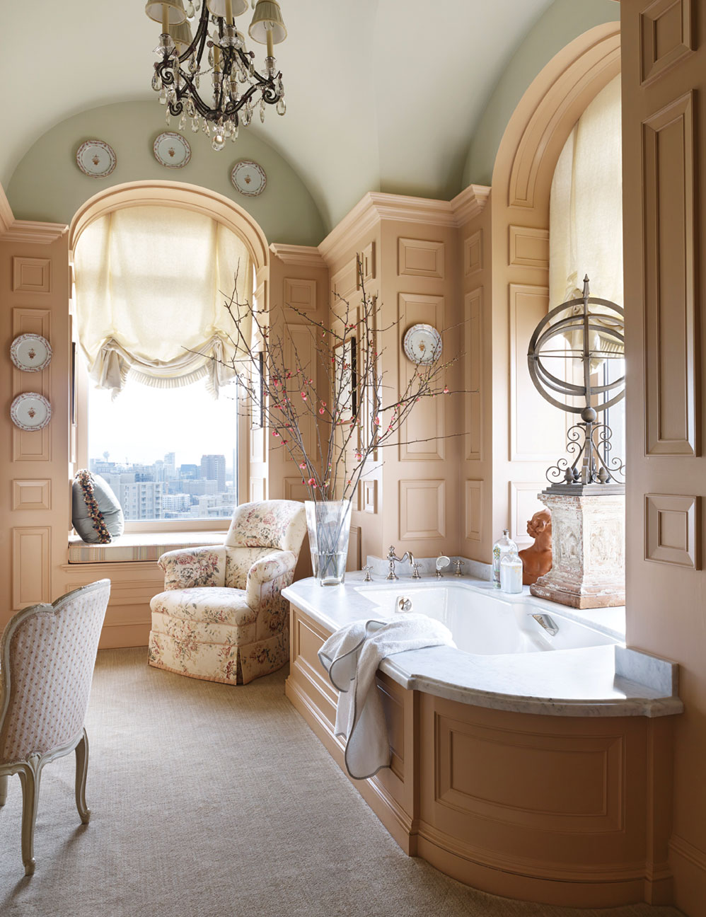 Other luxury bathroom decor shown hear includes a crystal chandelier, a cushioned window seat, tall arched windows and ceilings, and comfortable chairs.