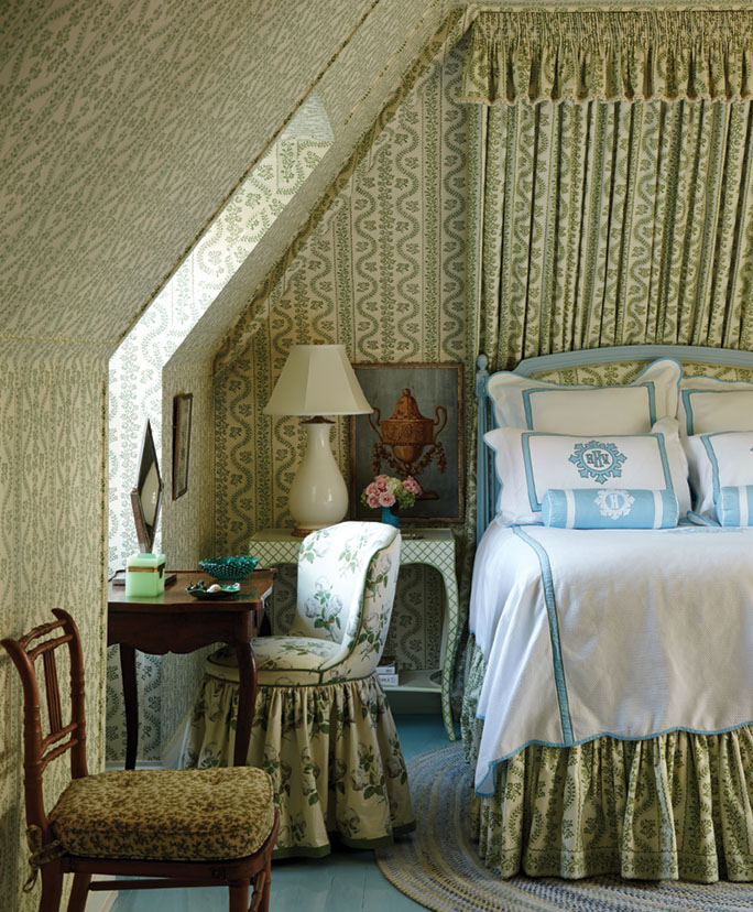 Behind the blue painted bedframe, a gathered, floor-to-ceiling fabric panel with valance displays the same botanical, sage green print as the wallpaper that envelops the space. The bed skirt also features the ‘Dolly’ Sister Parish print, while a skirted upholstered chair at the small antique desk by the window features a complementary floral pattern. Fresh white bedding is trimmed in a robin’s egg blue.