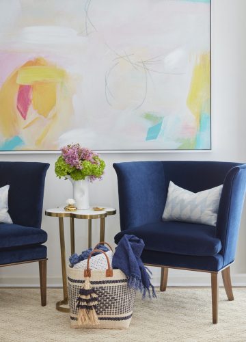 Two deep blue wing chairs flank a small side table with a brass base and stone top. On the wall hangs a very large, airy abstract painting, with a primarily white (or very pale) background and punches of bright yellow, pink, aqua and green.