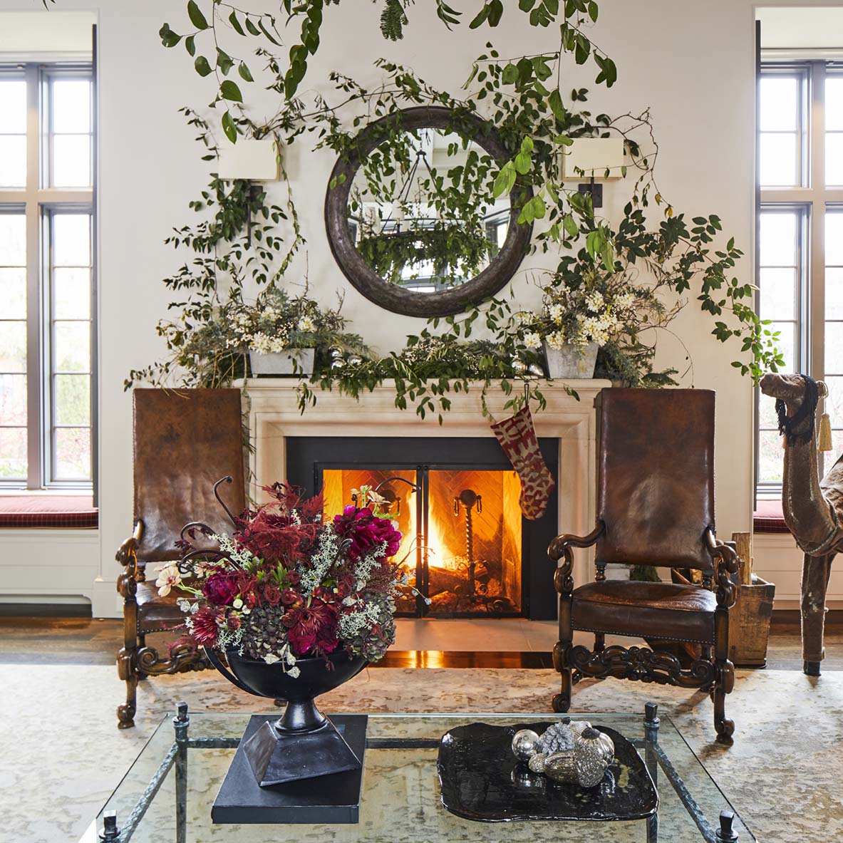 Two antique carved wood chairs upholstered in leather flank a fireplace decorated with greenery arrangement as if it were growing naturally on the mantel and wall.