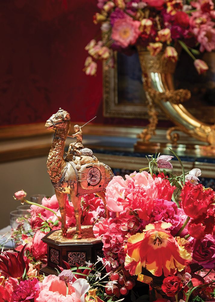 The lush floral arrangement features a range of pink and coral flowers, with touches of yellow, around a gilded sculpture of a woman riding a camel