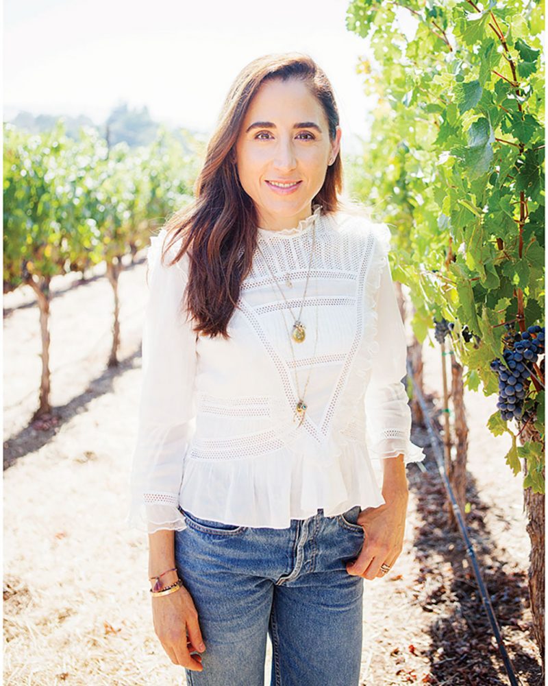  April Gargiulo stands in a vineyard wearing a white 3/4-sleeve top and jeans