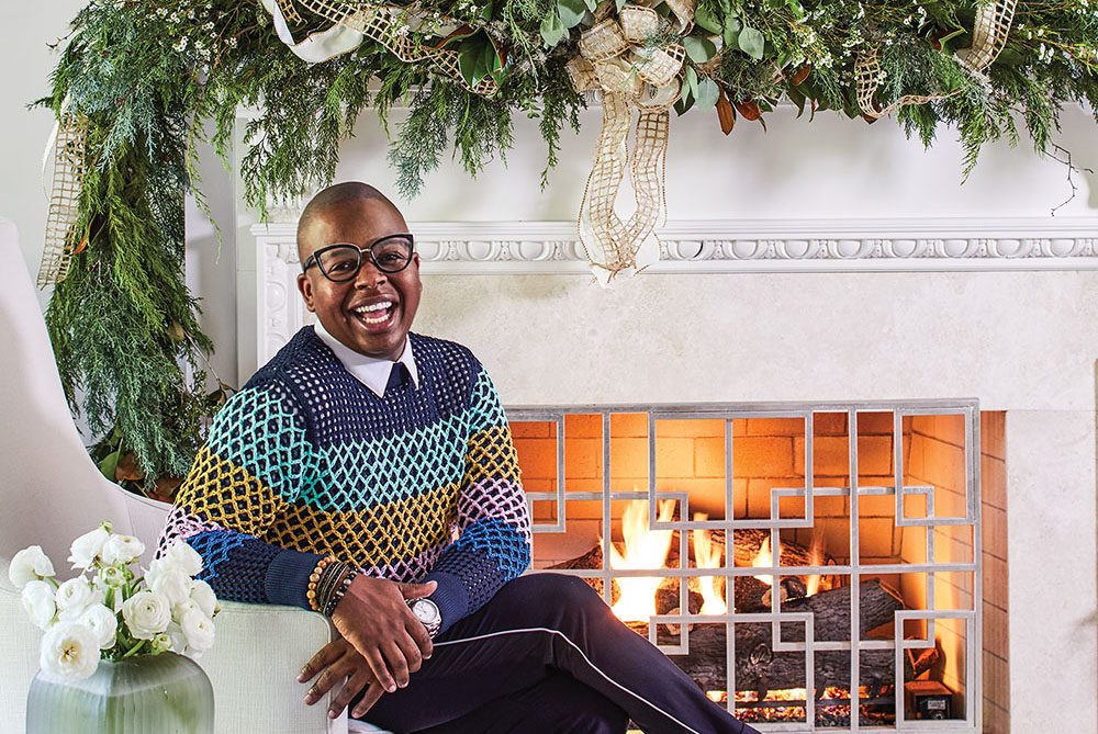 Atlanta floral designer Canaan Marshall, wearing a stylish crochet sweater, tailored dark pants a thin light strip along the outer seam, and loafers. The mantel is elaborately decorated with greenery and gold ribbon for the holidays.