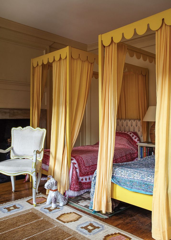 The room, as seen in 'Near & Far' by Lisa Fine, features two twin beds with scalloped-edged canopy frames painted yellow. Yellow fabric panels hang from the four corners of each canopy. One bed wears a blue and white patterned quilt; the other wears a red and white patterned quilt. A white French antique chair sits at the foot of one bed, next to a small dog statue. An area rug features subtle stripes in brown, framed by a natural-white border that displays geometric designs in soft blues and neutrals.