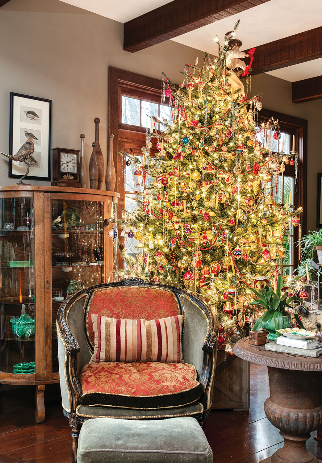 Living room vignette with an antique upholstered chair, antique wood and glass display cabinet, and a blue spruce covered in ornaments and white holiday lights