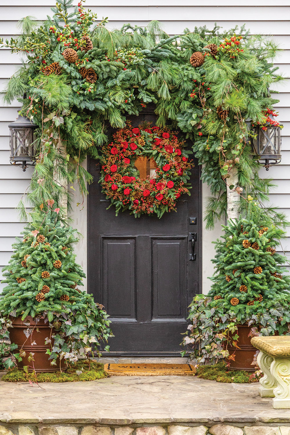 Front door holiday decor, festooned with greenery and red berries, flanked by evergreens, with huge wreath on door. 