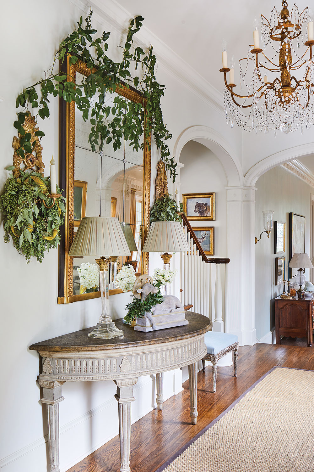 The painted foyer's architecture features high ceilings and gracious arches. Jane Schwab decorated it with a gilded chandelier adorned with delicate crystal swags, a simple sea grass or sisal rug, a semi-circle entry table, and a large gilded antique complemented by Italian sconces on either side.