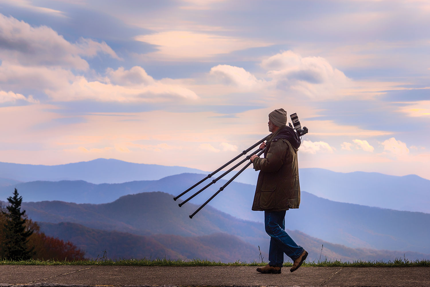 Sam Stapleton dressed in winter gear carries his tripod and camera as he walks along as sidewalk with a mountainous view in the background.