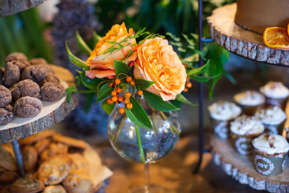A small vase of apricot-colored roses, berries, delicate foliage sits among trays of assorted cookies and cupcakes at the holiday open house