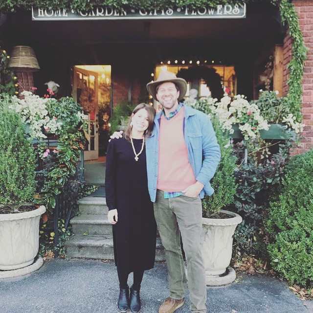Libby Endry, wearing a long casual black dress and gold necklace, with James Farmer, dressed in chinos, a peach sweater over a plaid button, light blue jacket, and brimmed hat, stand in front of the Gardener's Cottage front porch, surrounded by a lush container garden