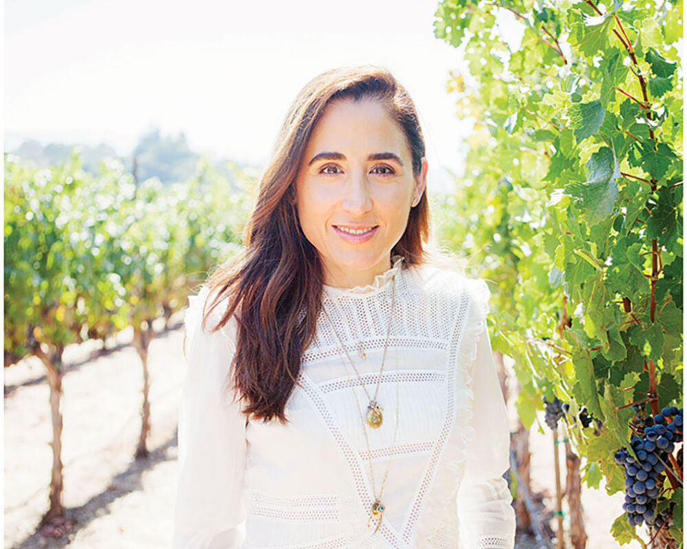 April Gargiulo, founder of Vintner's Daughter skin care, stands in a vineyard wearing a white 3/4-sleeve top and jeans