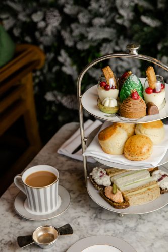 A tiered tray of sweets and savory finger sandwiches sits beside a cup of hot tea on a marble table at Dalloway Terrace in London
