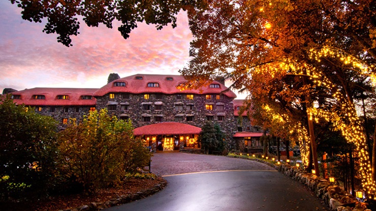 Best hotels in Asheville, NC: A view of the stone facade of The Grove Park Inn against a pink sunset. Twinkling tree lights follow the curve of the driveway, leading to the hotel entrance.