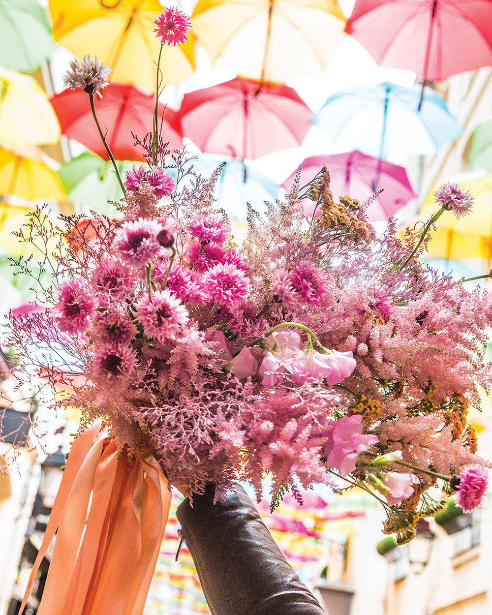 A hand holds lush pink bouquet tied with streaming peach ribbon against a colorful sky of suspended umbrellas
