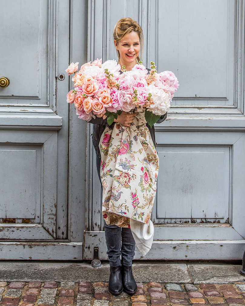 Parisian florist Catherine Muller stands on a brick-lined street in front of towering wooden double doors painted pale blue. She looks slightly away from camera and smiles, holding a profuse French bouquet of pink and white blooms, with the stems wrapped in a trailing floral fabric